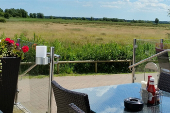 An outside pub beer garden, showing reflective glass tables/black wicker chairs on a concreted area in the foreground, with flat, light-brown fields in the distance on a cloudy day 