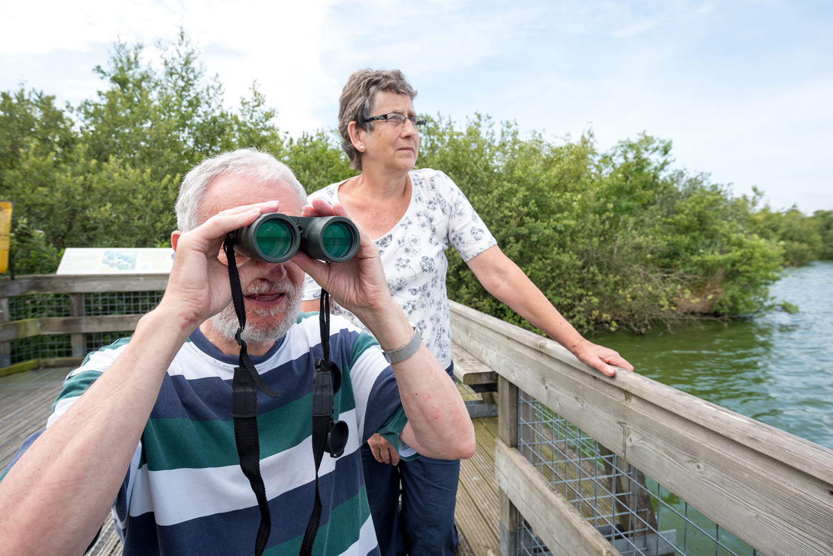 Two people stood on a wooden boardwalk birdwatching, holding binoculars