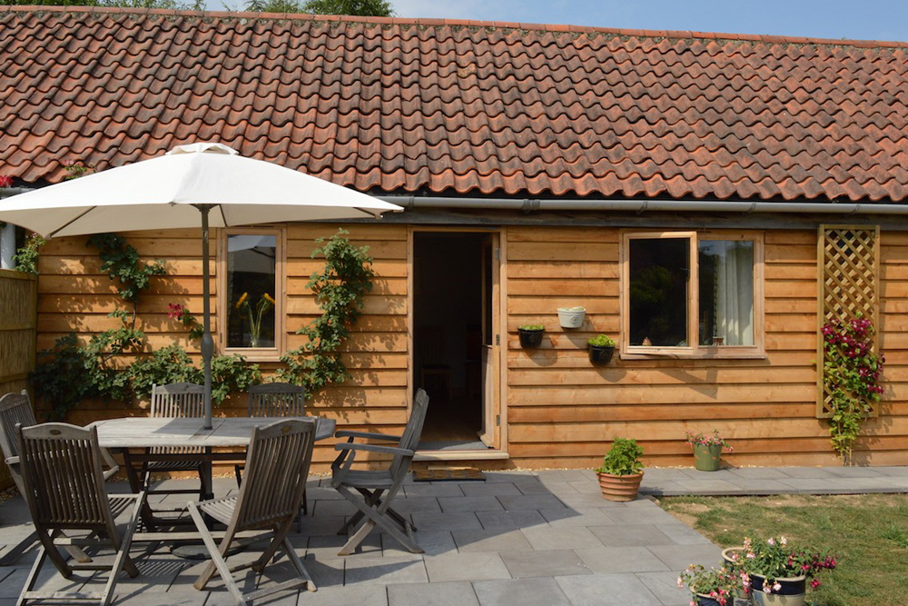 Exterior of a brown wood-cladded barn, with a grey path in the foreground and black umbrella parasol and seating area to the left