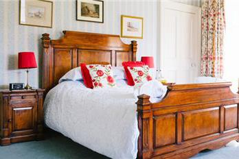 A large brown wooden double bed with white walls and white sheets. A red pillow is on the bed and a red lamp is on a wooden bed stand next to it