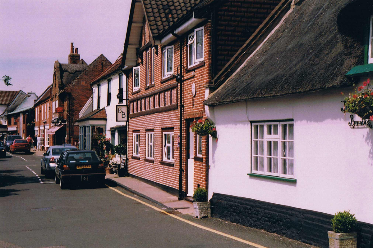 A street of thatched cottages, with a street stretching in to the distance, a white cottage is in the foreground and a red-bricked cottage is next door, other historic buildings are next to the cottage, and cars are parked outside