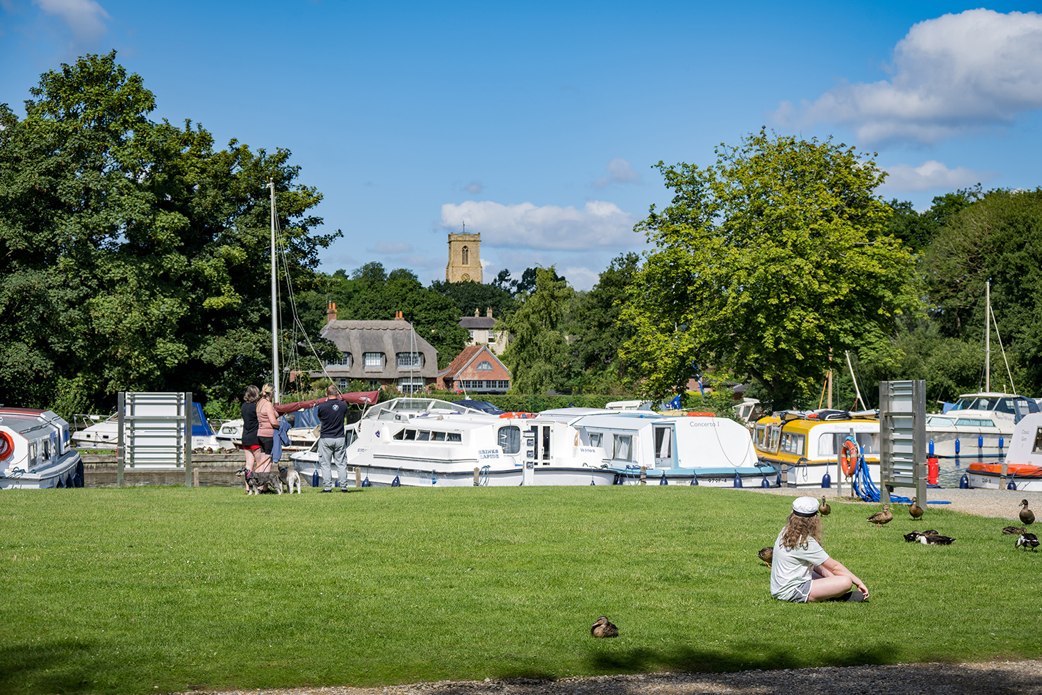 People sitting on the green at Ranworth Staithe on a sunny day with moored boats and St Helen's Church visible in the background