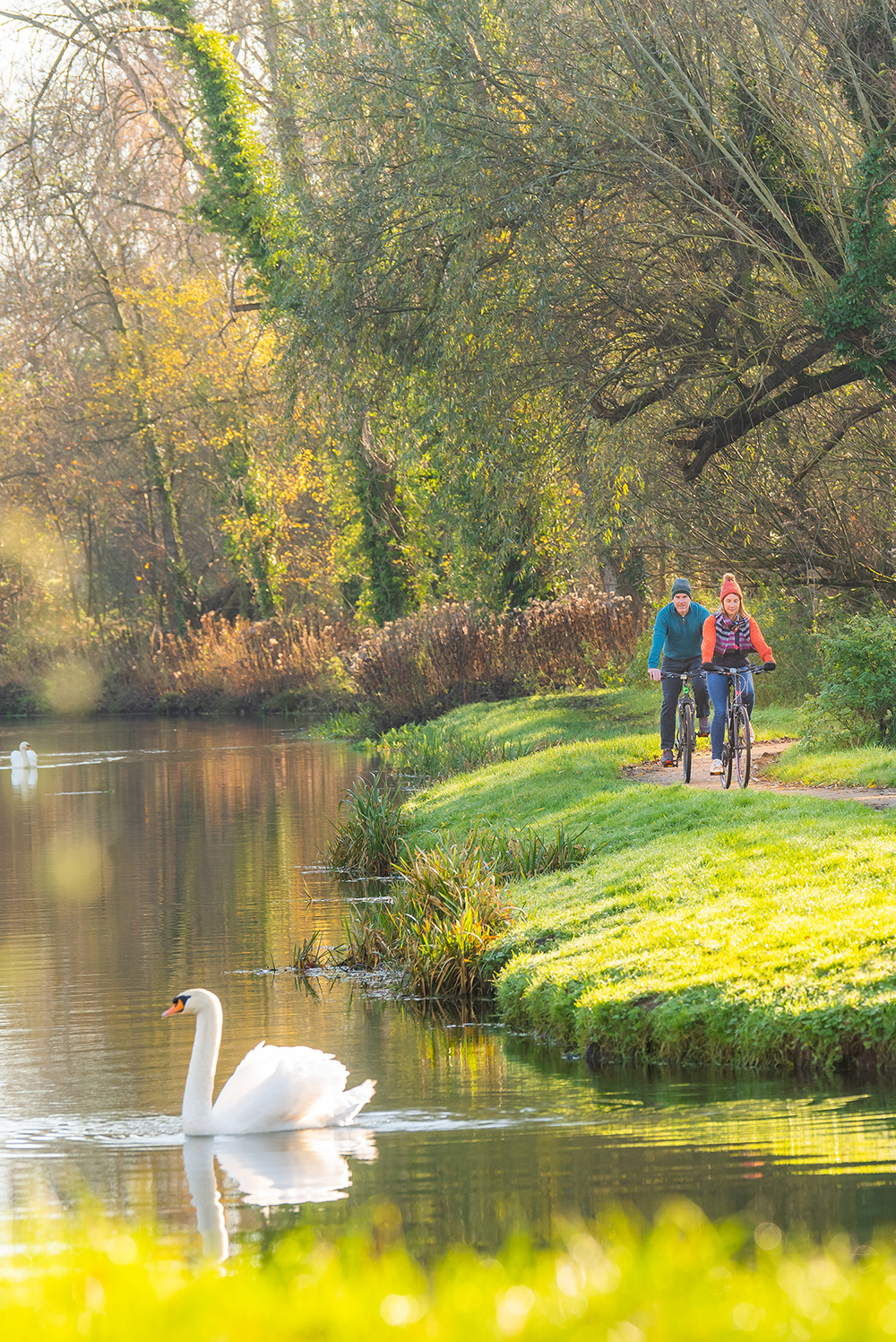 Two people wearing helmets ride bicycles along a dirt path beside a calm river on a sunny day, with blue skies and scattered clouds overhead. The path is surrounded by grassy fields and bare trees in the distance.