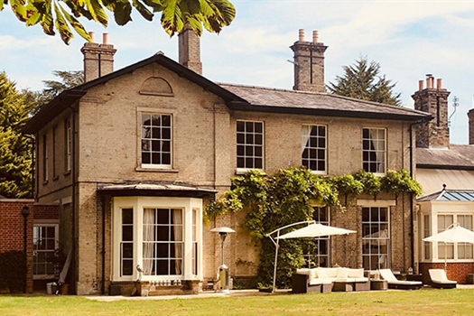The frontage of a large, grand grey Georgian hall, a green climbing bush is situated around the door of the property and there is a white seating area with umbrellas just in front