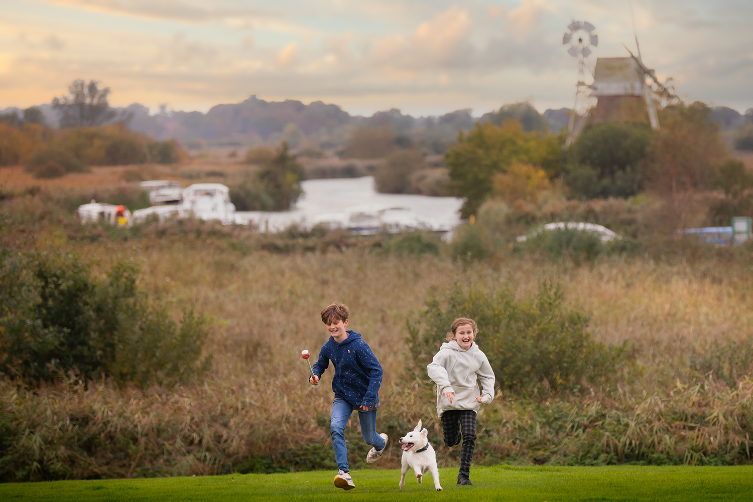 Two children and a dog run and play on a grassy surface with the a broads mill visible in the background.