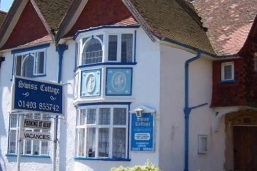 A close up view of a white rendered cottage, a blue sign says 'Swiss Cottage' in script writing, with a phone number underneath reading '01493 866742', drain pipes to the right of the frontage are blue in colour and there are ornate blue patterns underneath the upstairs windows