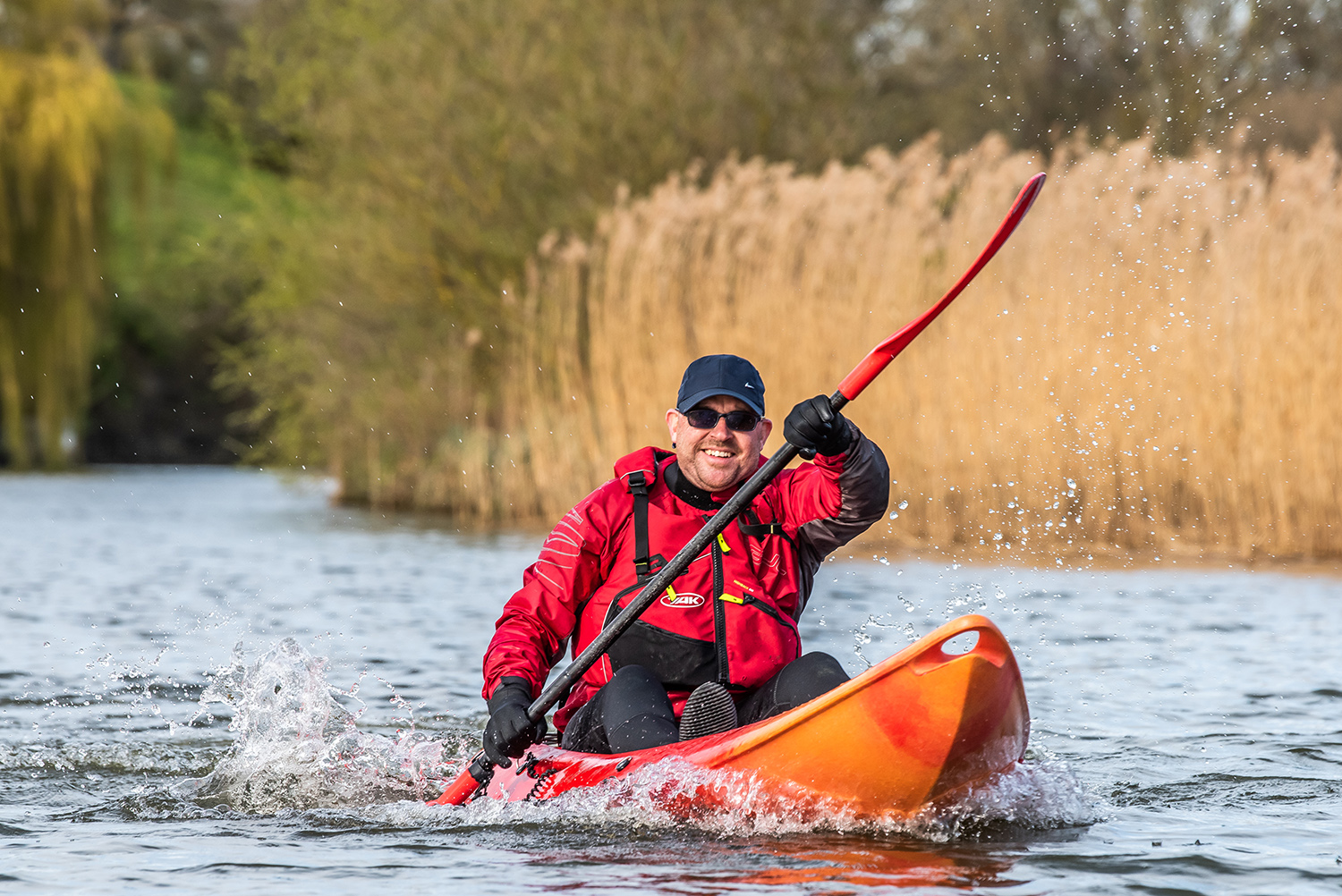 A man in a red jacket, black gloves, and sunglasses smiles while paddling a bright orange kayak on a river, splashing water as he moves past reeds and trees.