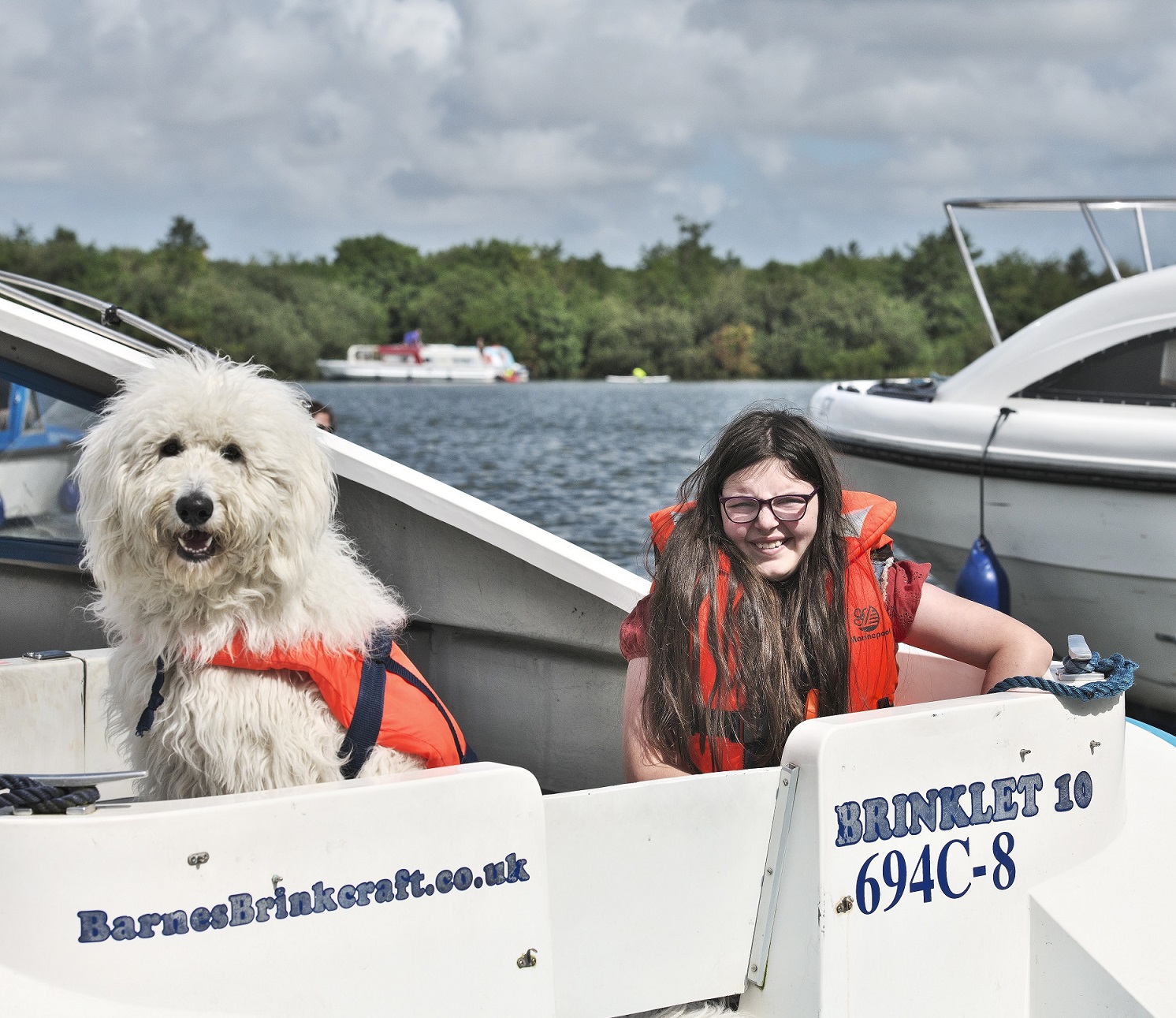 A young girl with brown hair and glasses is seated in the boat.  A small, white fluffy dog is also seated in the boat. Both the dog and girl are wearing life jackets. The background includes a shoreline with trees, and the sky is a light overcast grey.