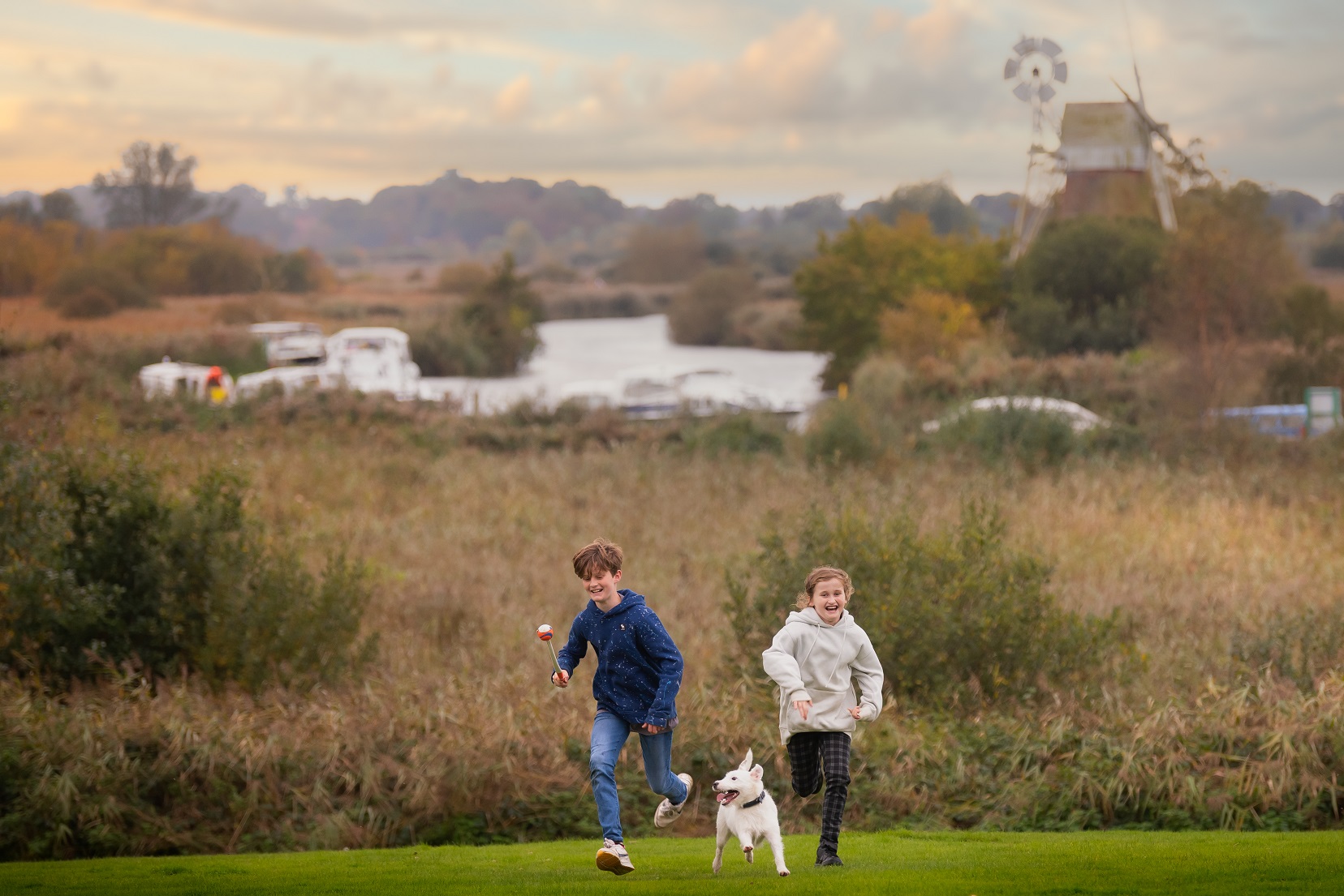 Two children and a white dog running on green grass with How Hill Nature Reserve in the background.
