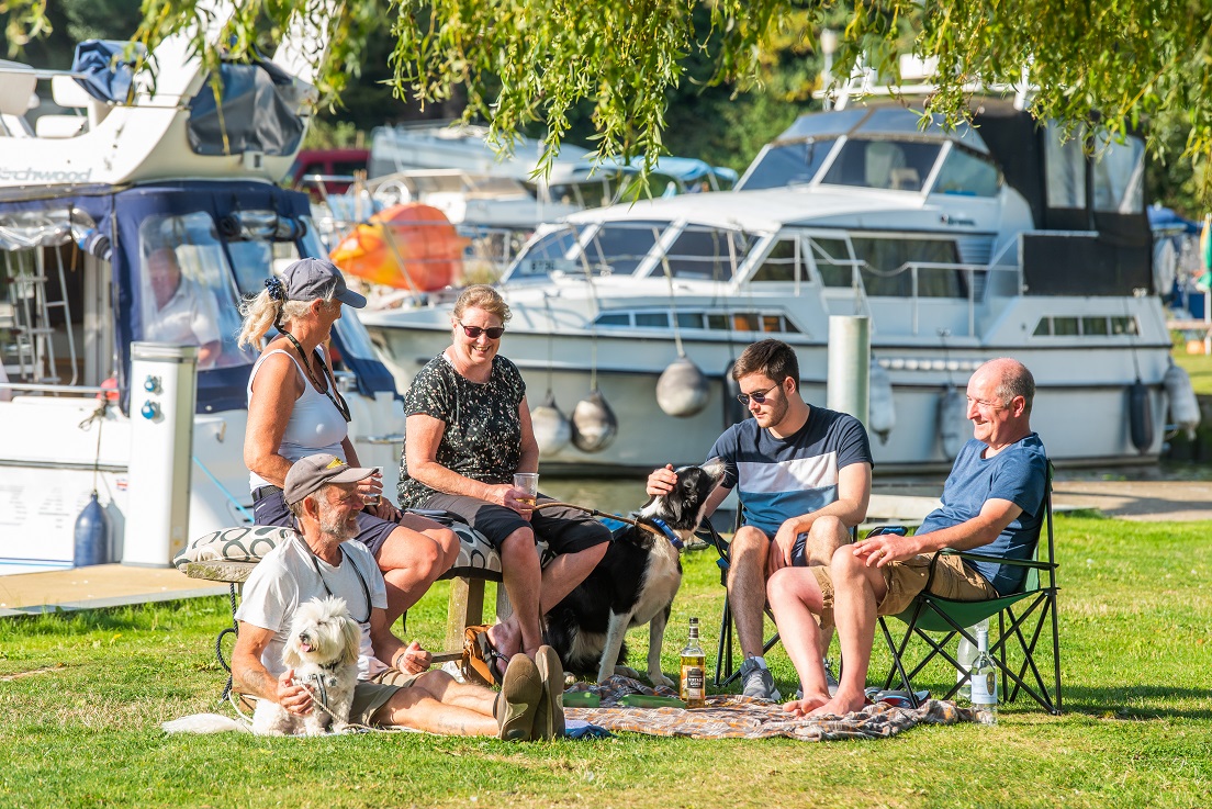 A group of people relaxing in a grassy area by a river. Two adults are sitting on foldable chairs. Another man is seated on a bench, and a woman is sitting next to him, both are wearing casual tops. There are several medium-sized dogs are present. There are several boats along the water and it is a sunny day. 