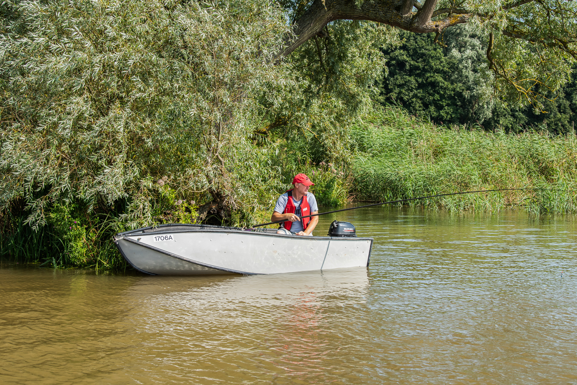 a man fishing from a boat in the broads surrounded by riverbank vegetation