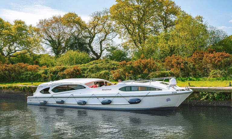 White Broadsman cruiser moored beside a tree-lined riverbank on the Norfolk Broads