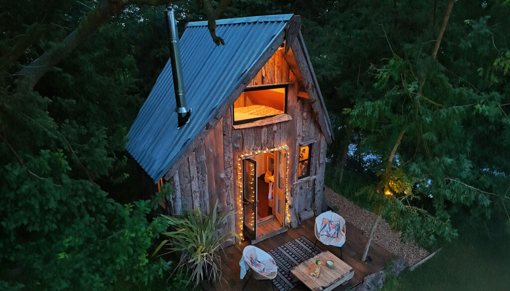 Evening outside of a rustic log cabin. A cosy light glows from inside