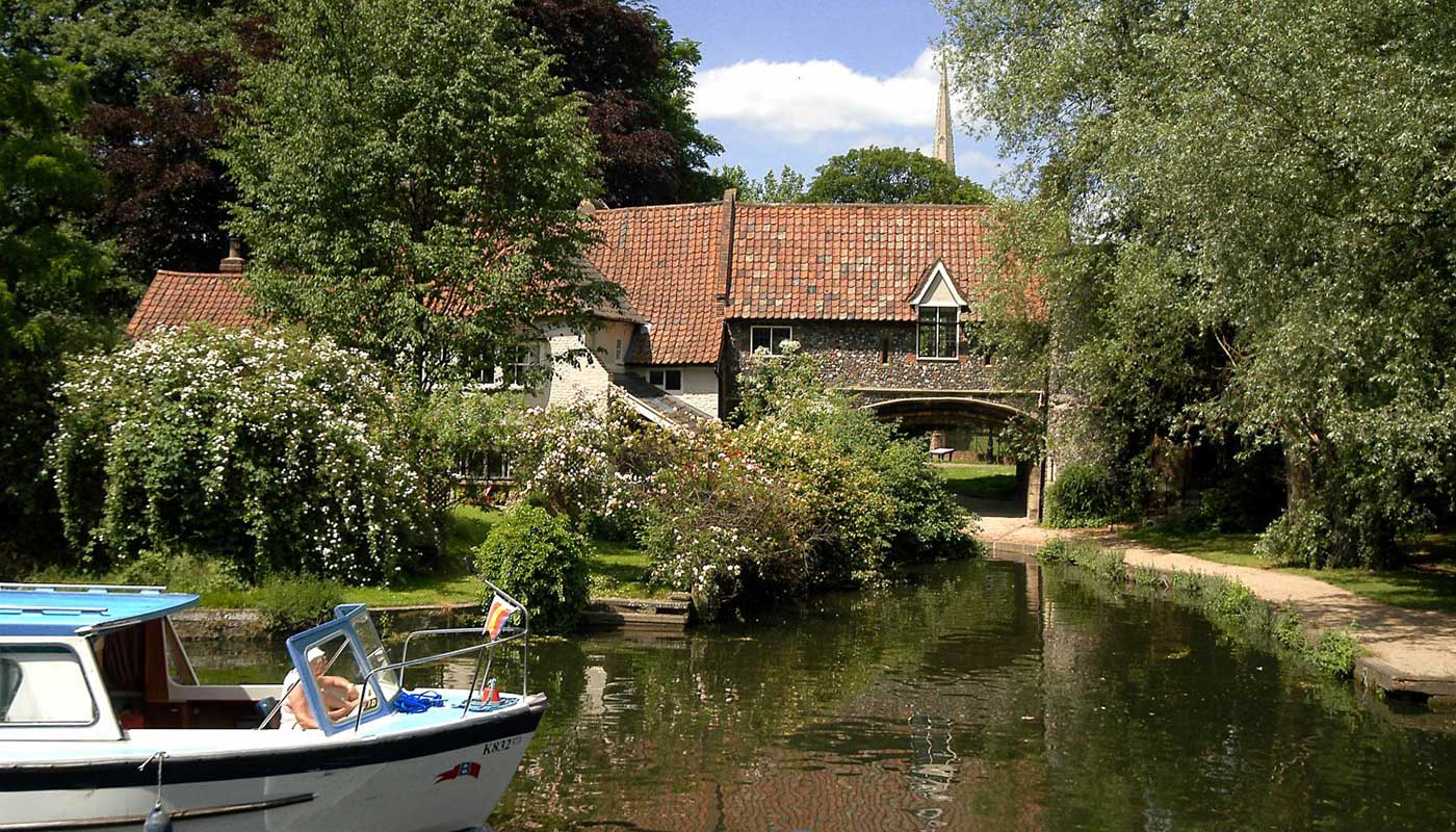 A dayboat passing Pulls Ferry in norwich, with the cathedral spire in the background.