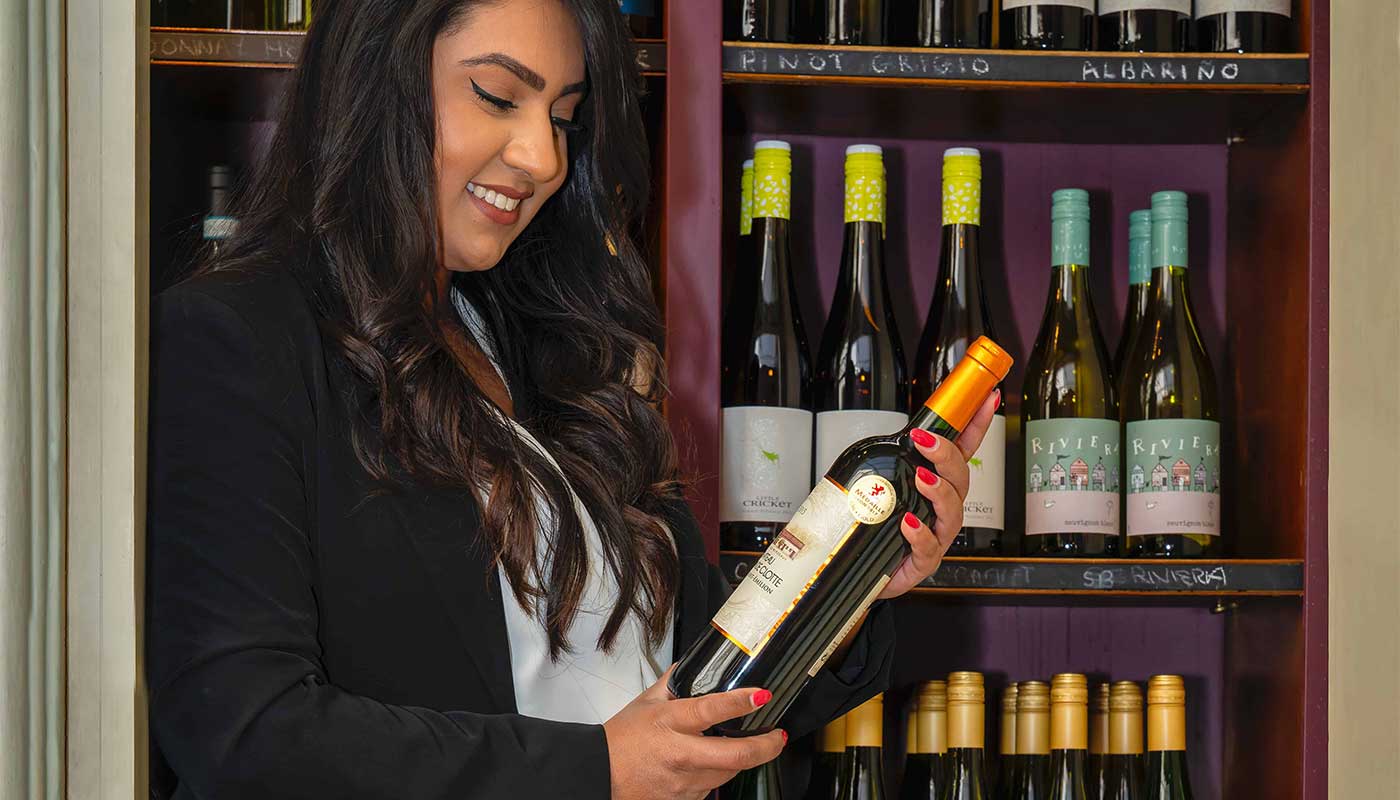 A lady stood in front of shelves of wine, looking at one of the bottles which she holds in her hand